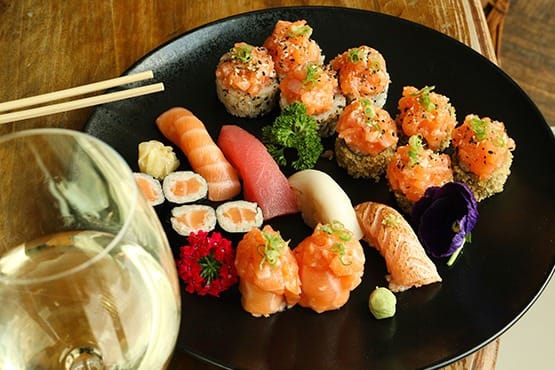 Um prato preto com sushi e sashimi variados, guarnecido com verduras e uma flor comestível, fica ao lado de uma taça de vinho branco e um par de pauzinhos de madeira.