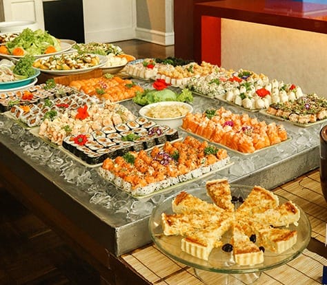 Uma mesa de bufê repleta de rolos de sushi variados, sashimi, saladas e guarnições, com um prato de vidro com fatias de torta em primeiro plano. Os pratos estão bem arrumados e decorados com verduras e flores.