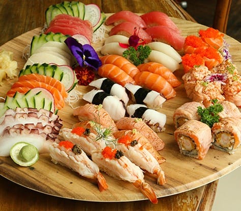 Um prato de madeira com uma variedade de sushi e sashimi, incluindo rolinhos, nigiri e fatias de peixe cru, guarnecido com pepino, wasabi, gengibre e flores comestíveis.