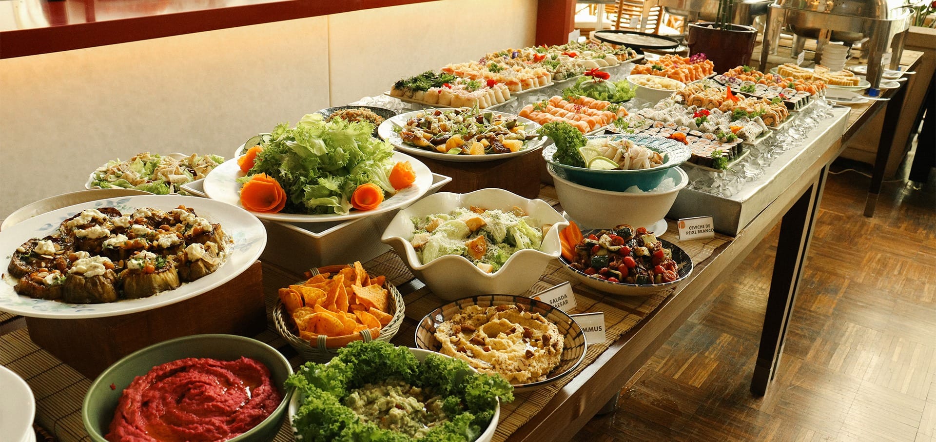 Uma mesa de bufê repleta de uma variedade de pratos, incluindo saladas, legumes, molhos, batatas fritas e aperitivos variados, dispostos ordenadamente em pratos e tigelas.