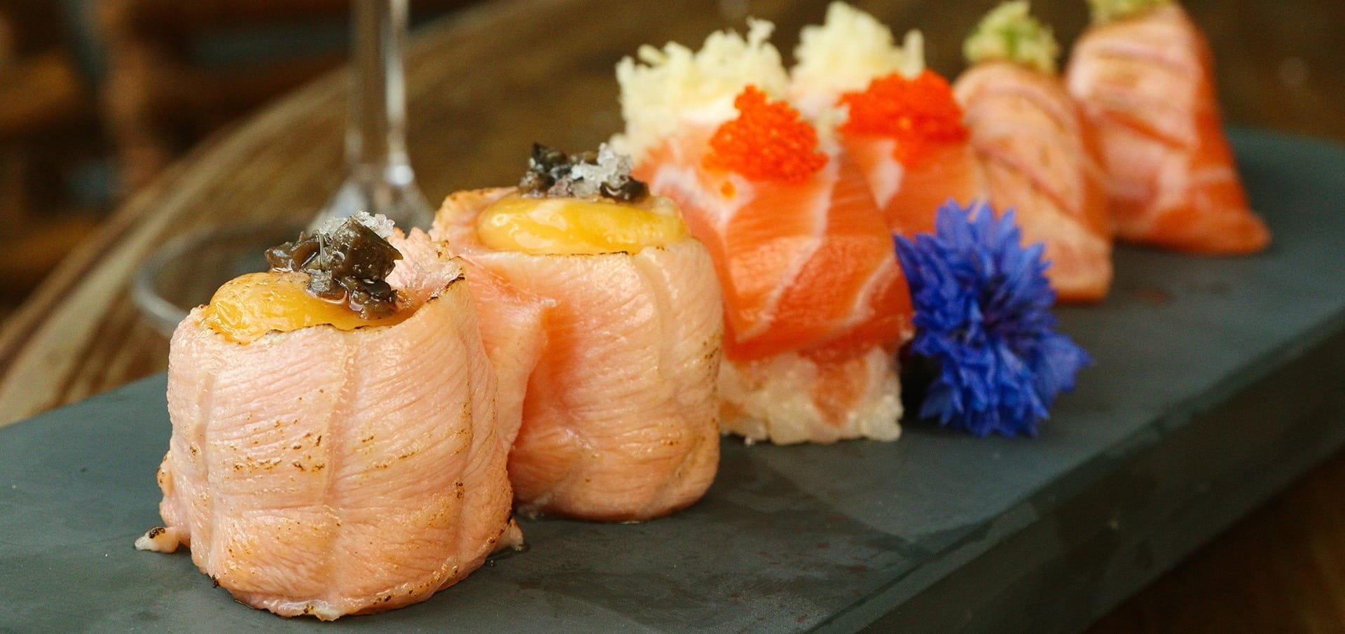 Um close-up de peças variadas de sushi em um prato de ardósia preta, incluindo salmão grelhado coberto com molho, nigiri com ovas brancas e laranja, guarnecido com rabanete picado e uma flor roxa comestível.