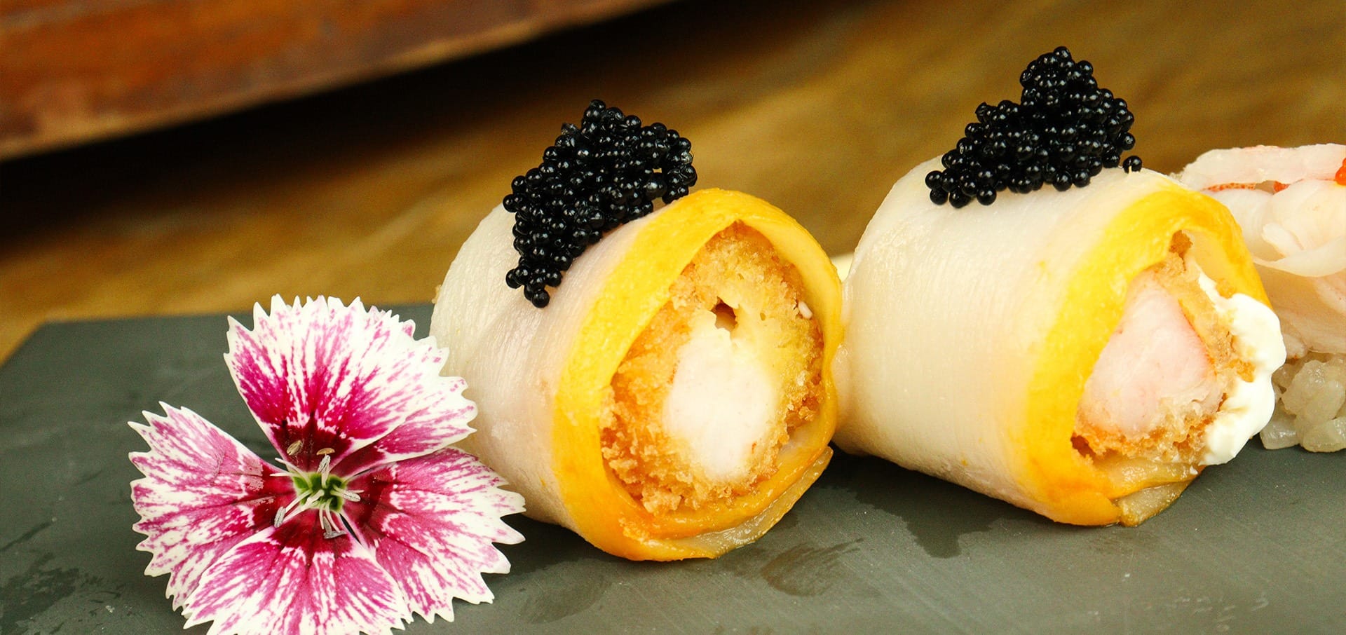 Dois pedaços de rolos de sushi cobertos com caviar preto ficam em um prato de ardósia, acompanhados por uma flor rosa e branca. O sushi parece ter um exterior dourado e um recheio cremoso em seu interior.