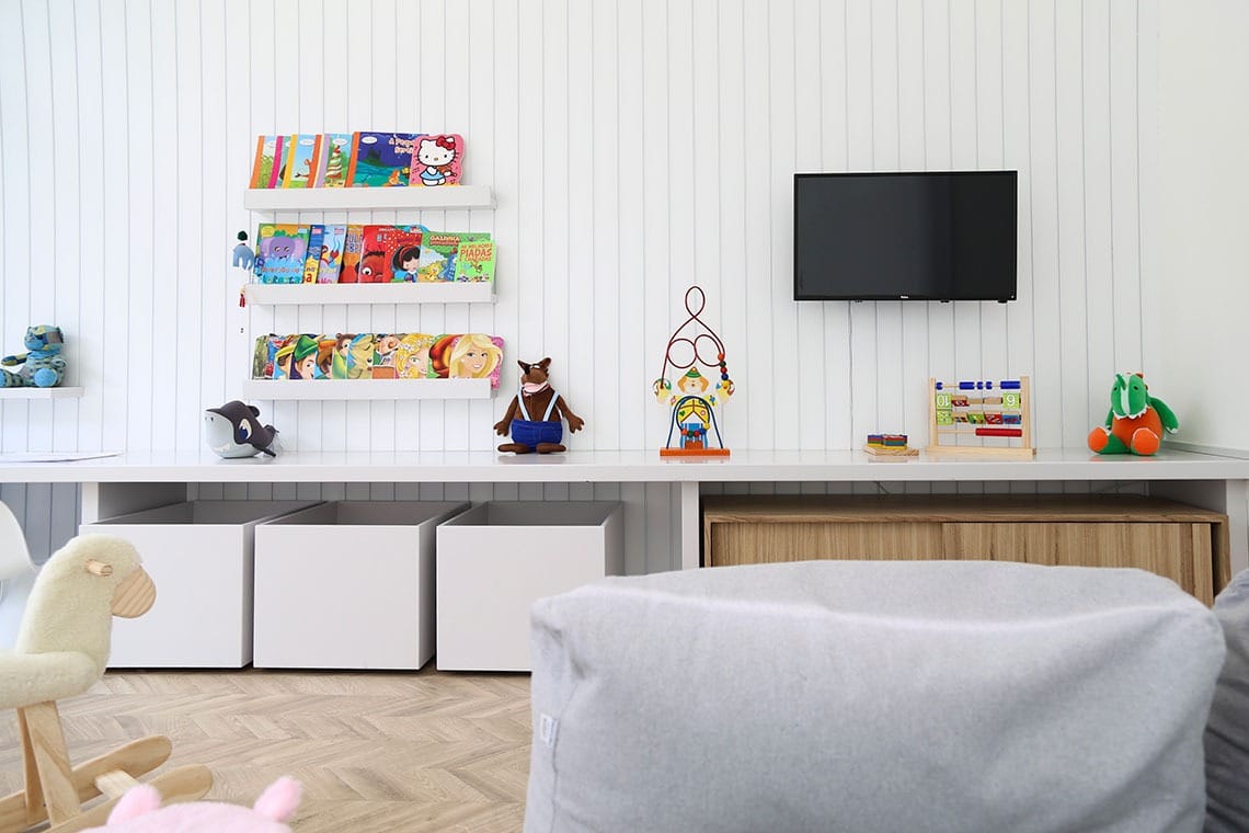 Uma sala de jogos infantil moderna e organizada, com paredes brancas, prateleiras de livros coloridos, brinquedos em um balcão comprido, uma TV na parede, caixas grandes de armazenamento, um cordeiro de balanço e uma cadeira cinza com saco de feijão em um piso de madeira.