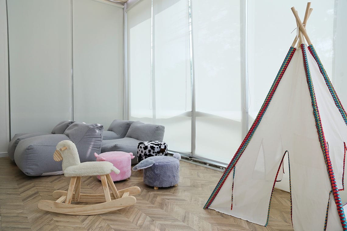 Uma sala de jogos moderna com cadeiras de saco de feijão cinza, um cavalo de balanço de madeira, banquetas macias em forma de animais e uma barraca de jogos infantis branca com detalhes coloridos em um piso de madeira em forma de espinha.