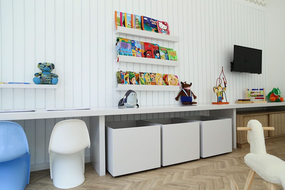 Uma sala de jogos infantil moderna e bem iluminada com paredes brancas, prateleiras de livros coloridos, vários bichos de pelúcia em uma mesa comprida, três caixas de armazenamento brancas embaixo e uma TV na parede. Há cadeiras azuis e brancas nas proximidades.