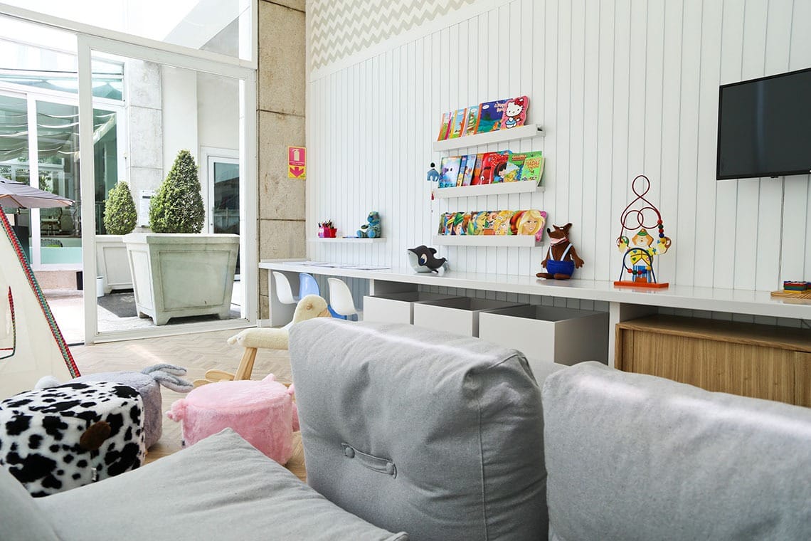 Uma moderna sala de jogos para crianças com assentos de pelúcia, banquetas com tema de animais, prateleiras com livros e brinquedos coloridos, uma TV na parede e grandes janelas que permitem a entrada de luz natural.